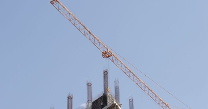 Orange tower crane working above an unfinished high-rise structure against a clear blue sky.