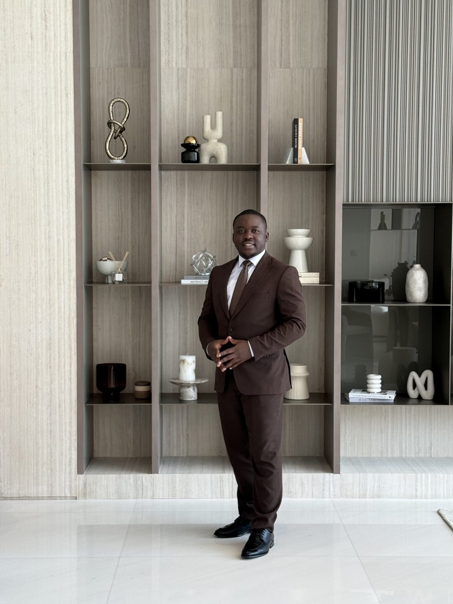 Hermann Tatang in a brown suit, standing in a finished Dubai show apartment in front of a travertine and dark-wood shelving wall.